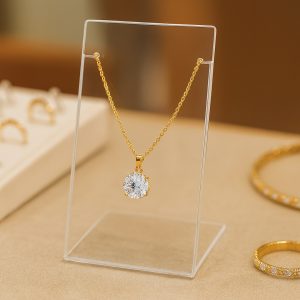 Clear acrylic necklace holder with side notches, displaying a gold necklace with gemstone pendant on a jewelry counter with rings and bracelets in the background